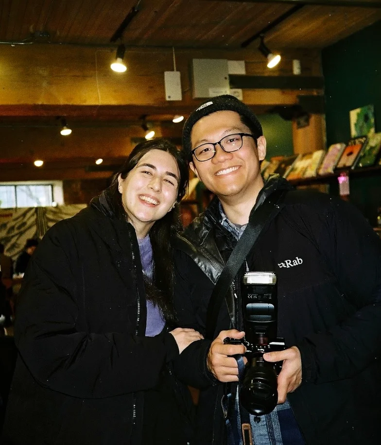 Two people smiling together, one holding a film camera, beside bookshelves.